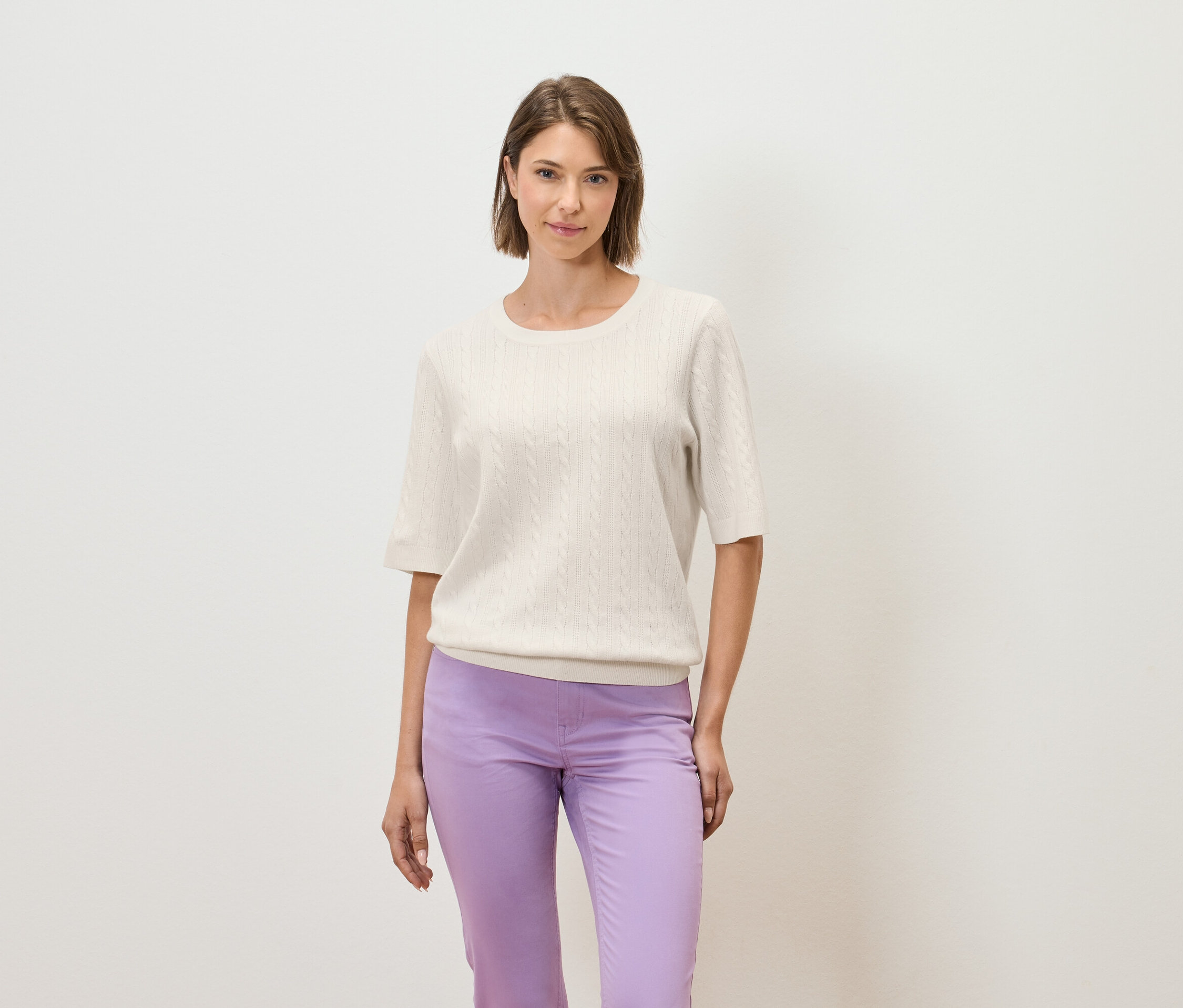 Eine Frau posiert vor einem hellen Hintergrund in einem cremefarbenen Kurzarmpullover und einer lila Hose.