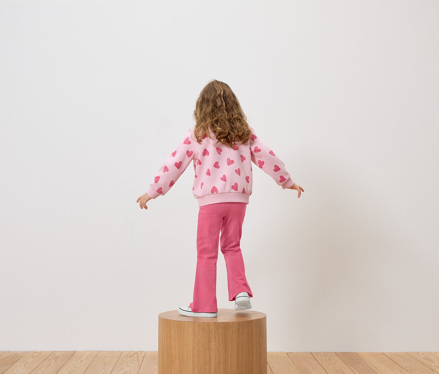 Mädchen steht mit dem Rücken zur Kamera auf einem Holzsockel, trägt einen rosa Pullover mit Herzchen und rosa Hosen.