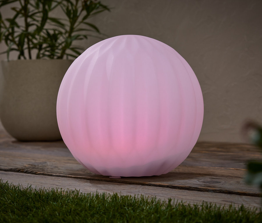 Eine pinke Kugel-Leuchte steht auf einem Holztisch, neben einem Topf mit einer Pflanze.