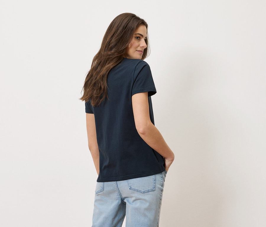 Frau mit dunklem T-Shirt und hellblauen Jeans steht vor einer weißen Wand.