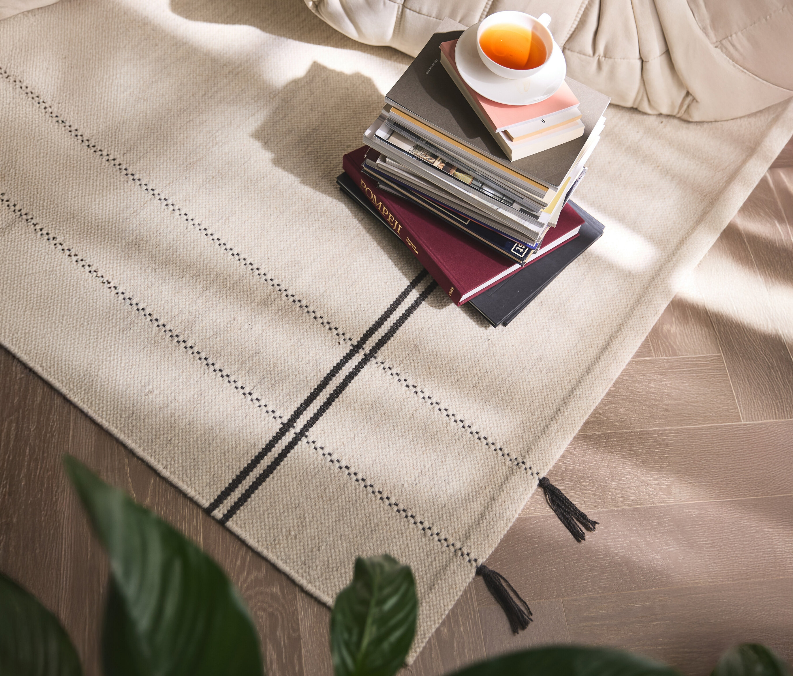 Ein beiger Teppich mit schwarzen Streifen und Fransen liegt auf einem Holzboden, darauf ein Bücherstapel mit einer Tasse Tee.