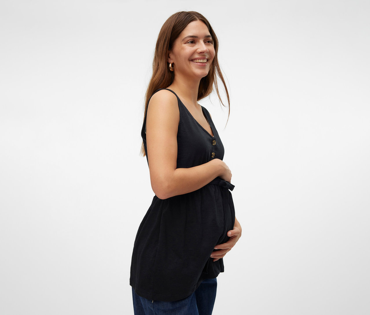Frau lächelt und hält ihren Bauch, während sie ein schwarzes MAMALICIOUS Stilltop »MLEVI LIA« und Jeans trägt.