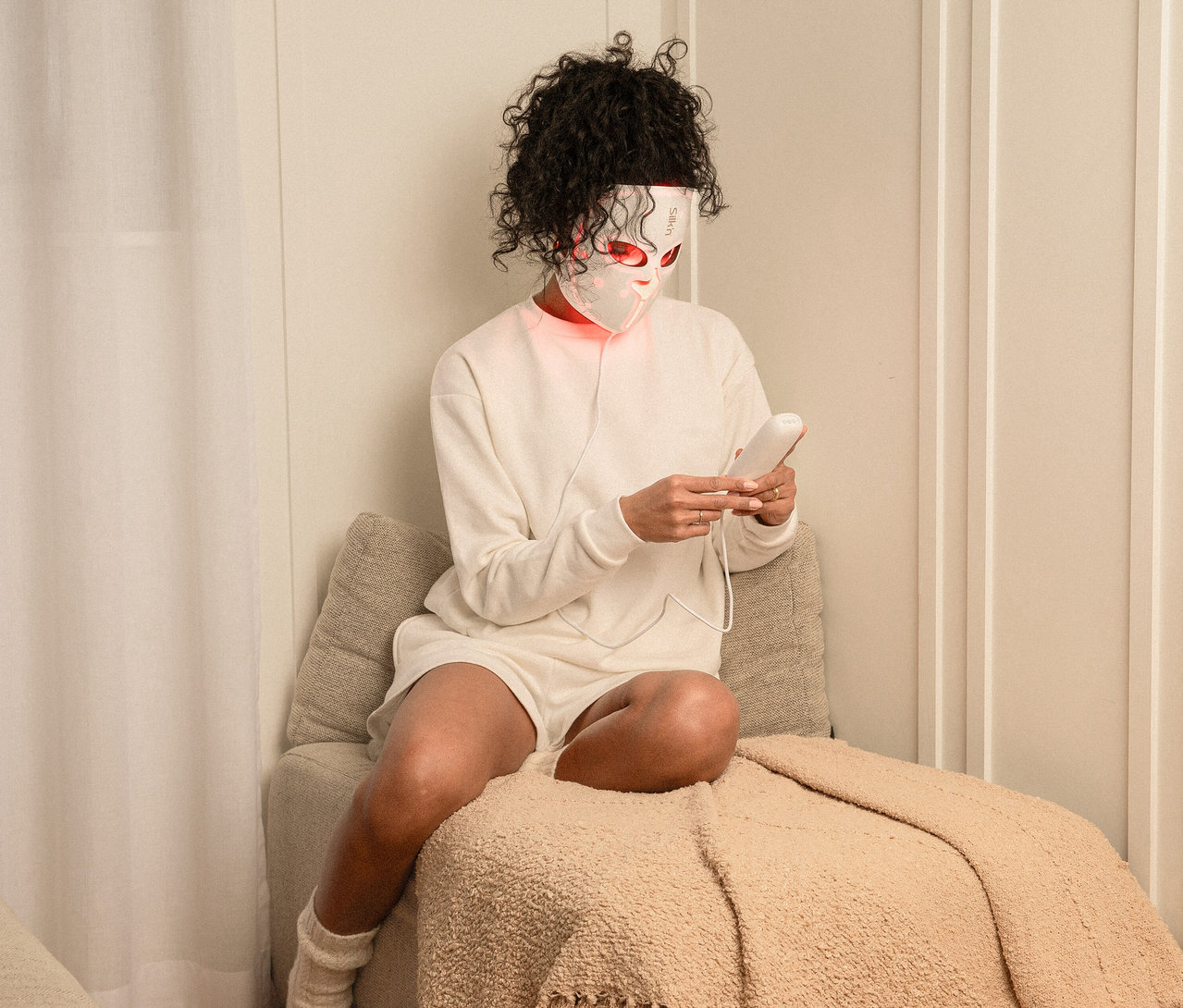 Frau sitzt mit Silk'n LED Gesichtsmaske mit EMS, hält Controller in den Händen.