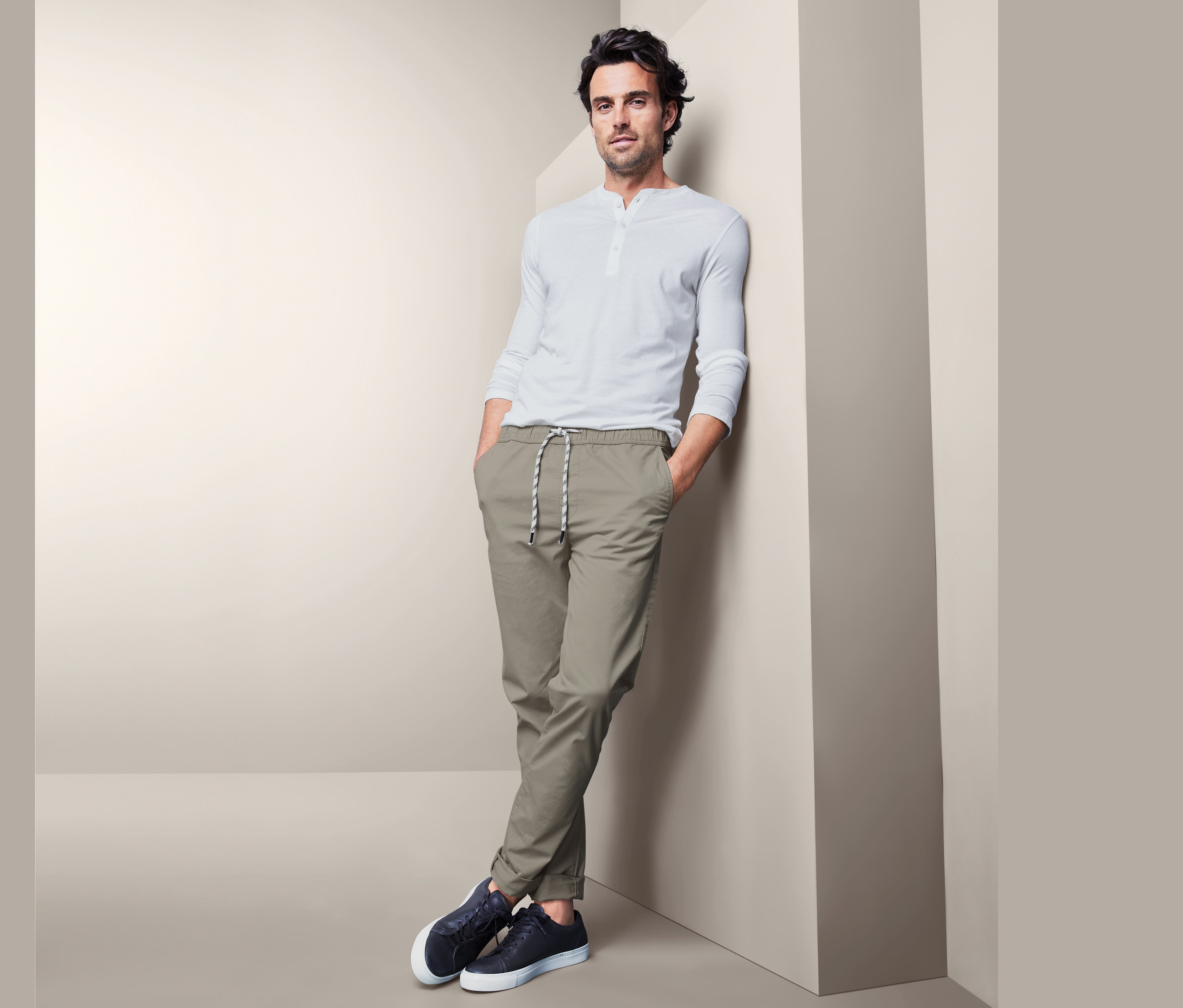 Mann lehnt an einer Wand, trägt eine Comfort-Chino, ein weißes T-Shirt und dunkelblaue Turnschuhe.