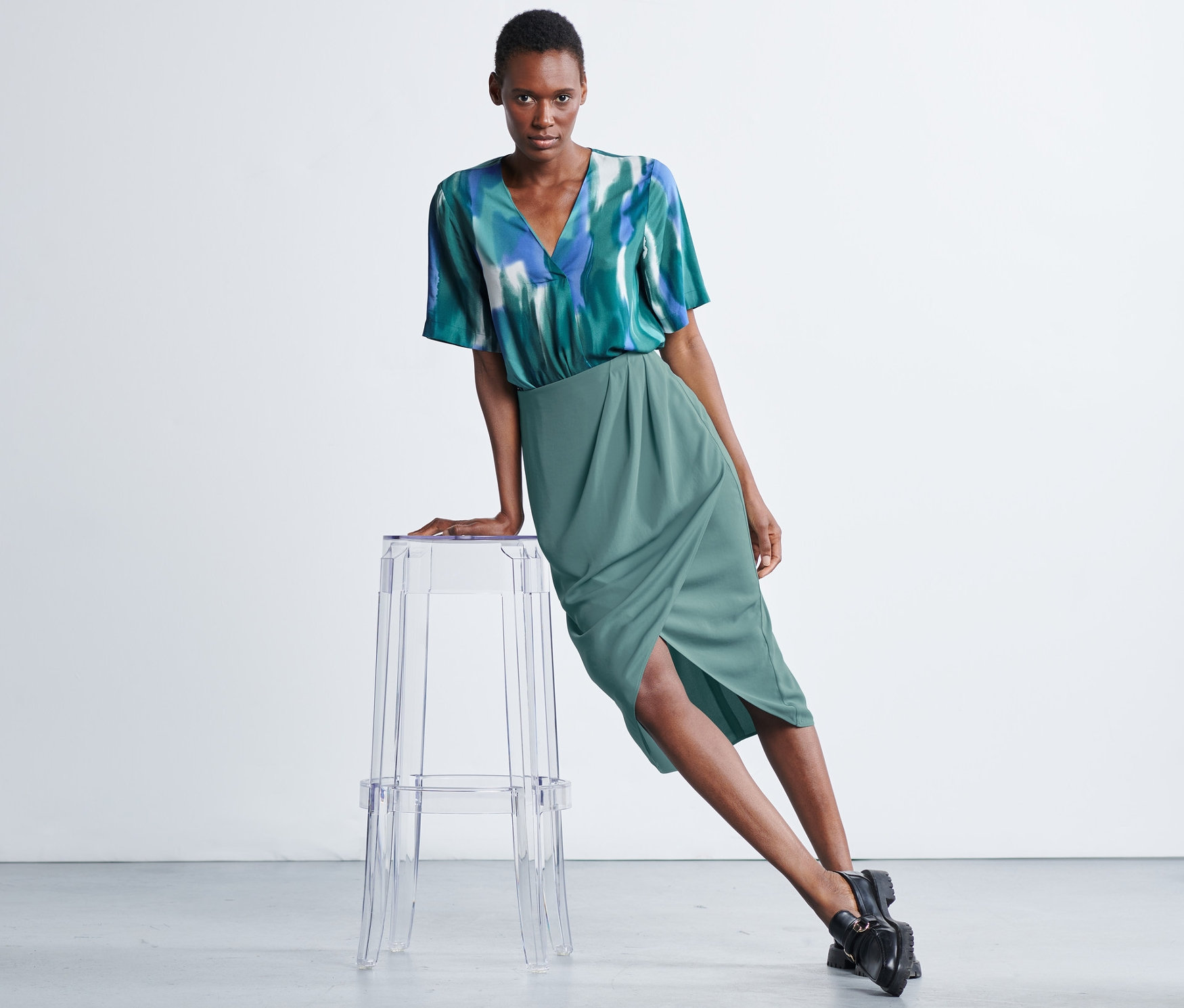 Frau, die sich an einen transparenten Stuhl lehnt, trägt einen Wickelrock und ein Top.
