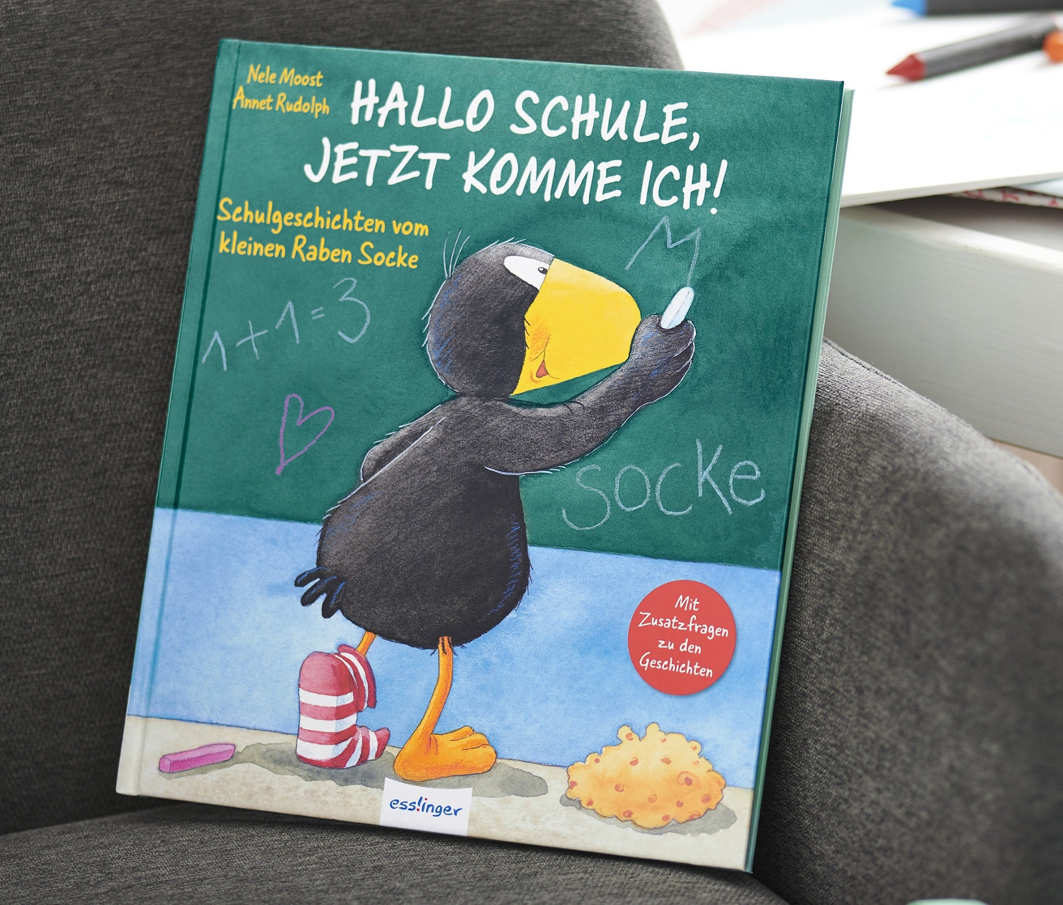 Auf einem grauen Sessel liegt das Buch »Hallo Schule, jetzt komme ich – Schulgeschichten vom kleinen Raben Socke«.