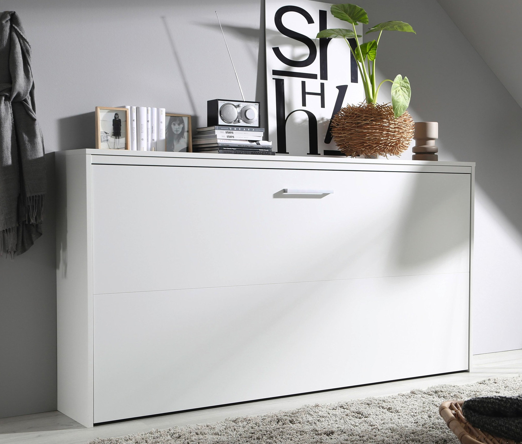 Ein weißes rauch Querklappbett »Albero« steht an der Wand, darauf Dekorationen. Im Vordergrund Teppich und Stuhl.