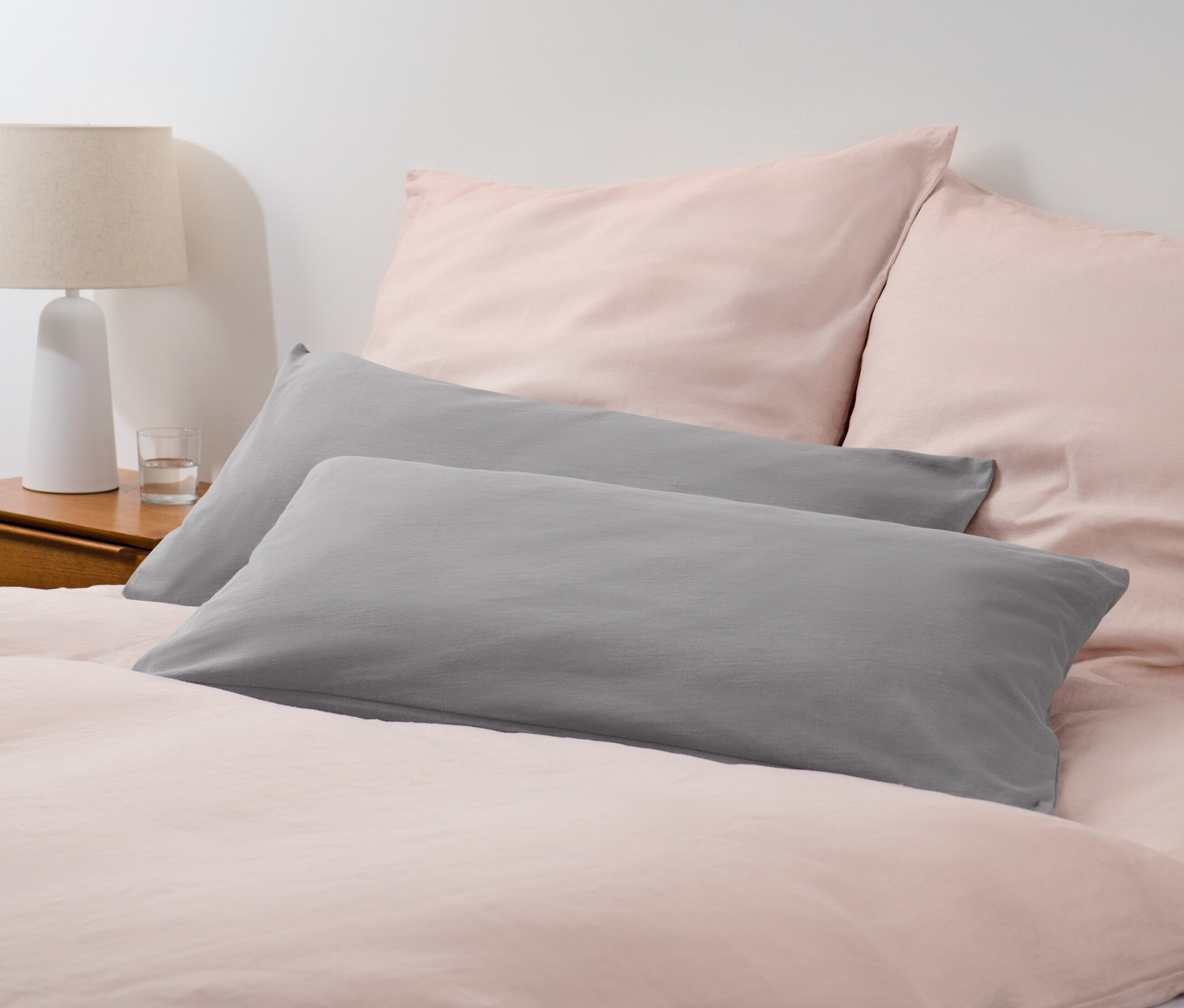 Zwei graue Jersey-Kopfkissenbezüge liegen auf einem Bett mit rosa Bettwäsche.