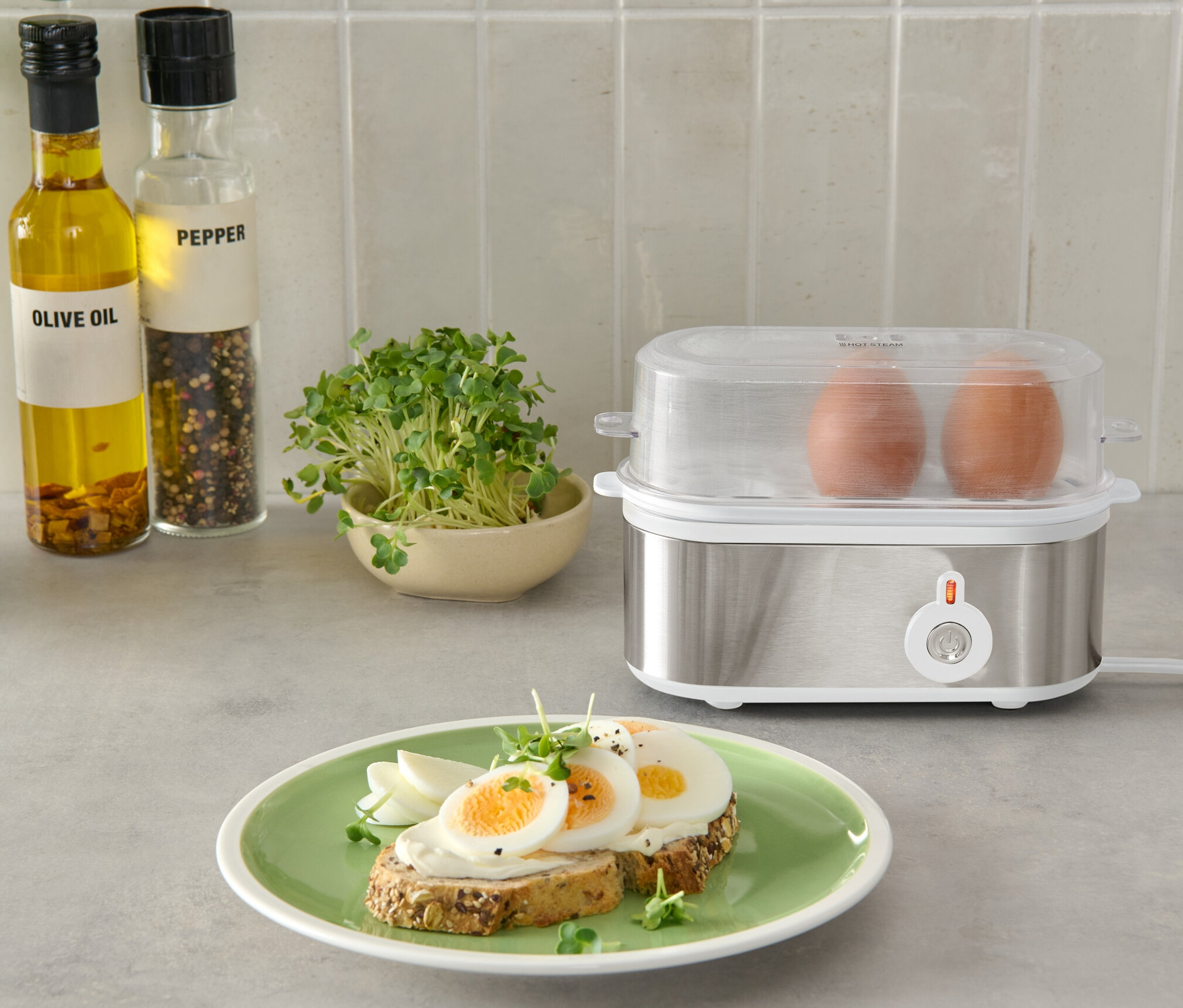 Zwei braune Eier in einem silbernen Eierkocher mit transparentem Deckel, neben einem Teller mit Brot und Eiern.