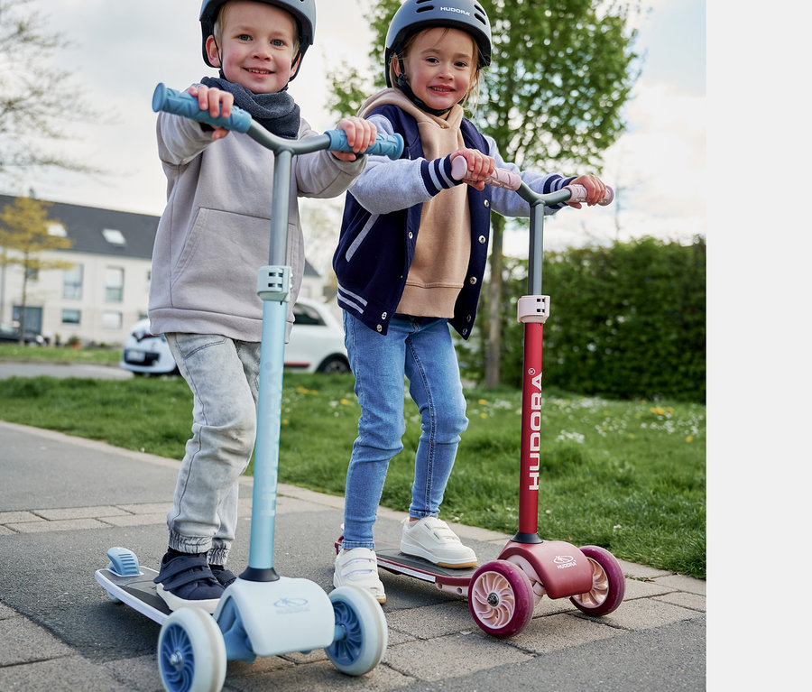 Zwei Kinder fahren mit einem bordeauxroten und einem blauen HUDORA Tri-Scooter auf dem Gehweg.