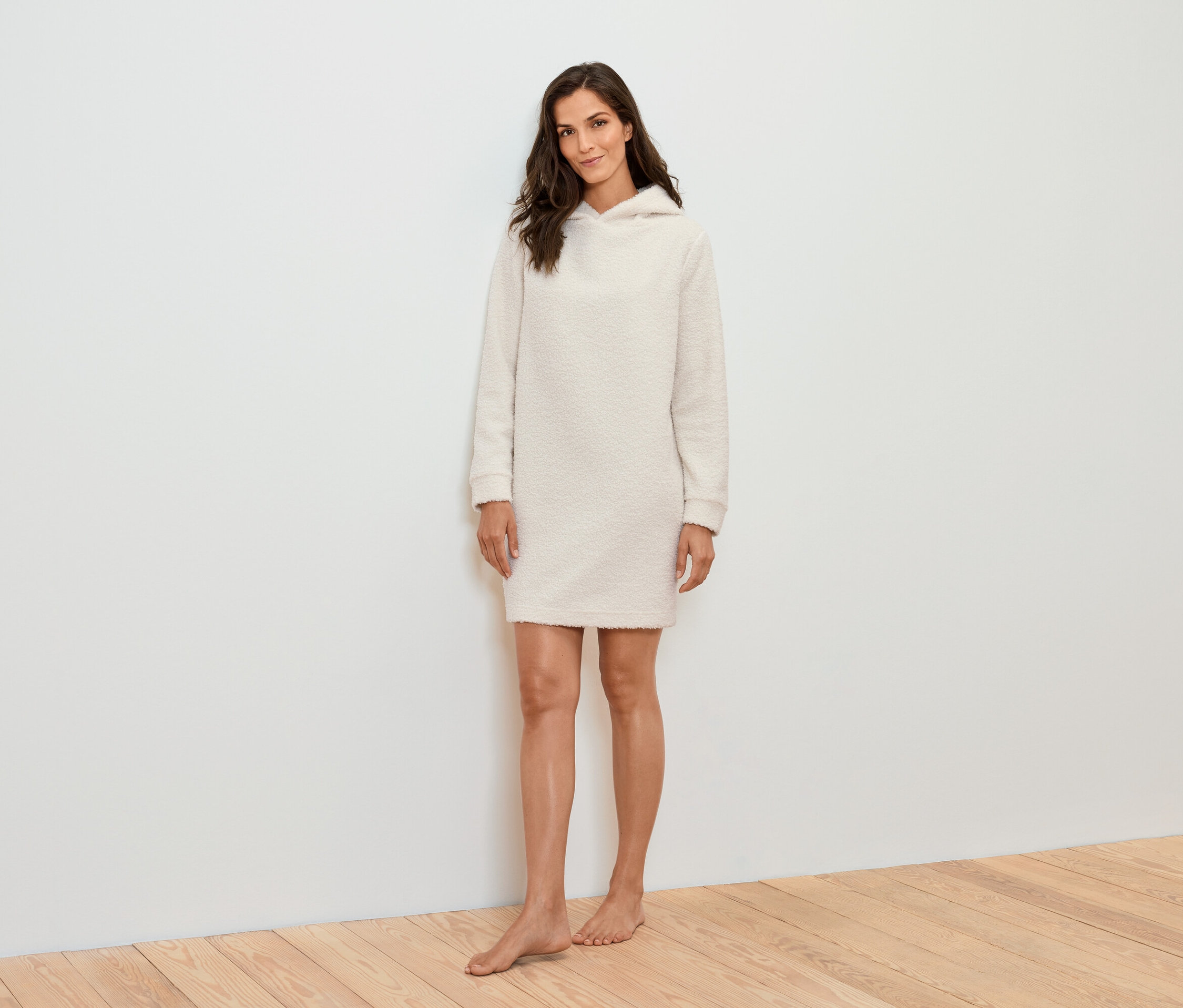 Eine Frau steht vor einer weißen Wand und trägt ein cremefarbenes Kapuzenkleid.