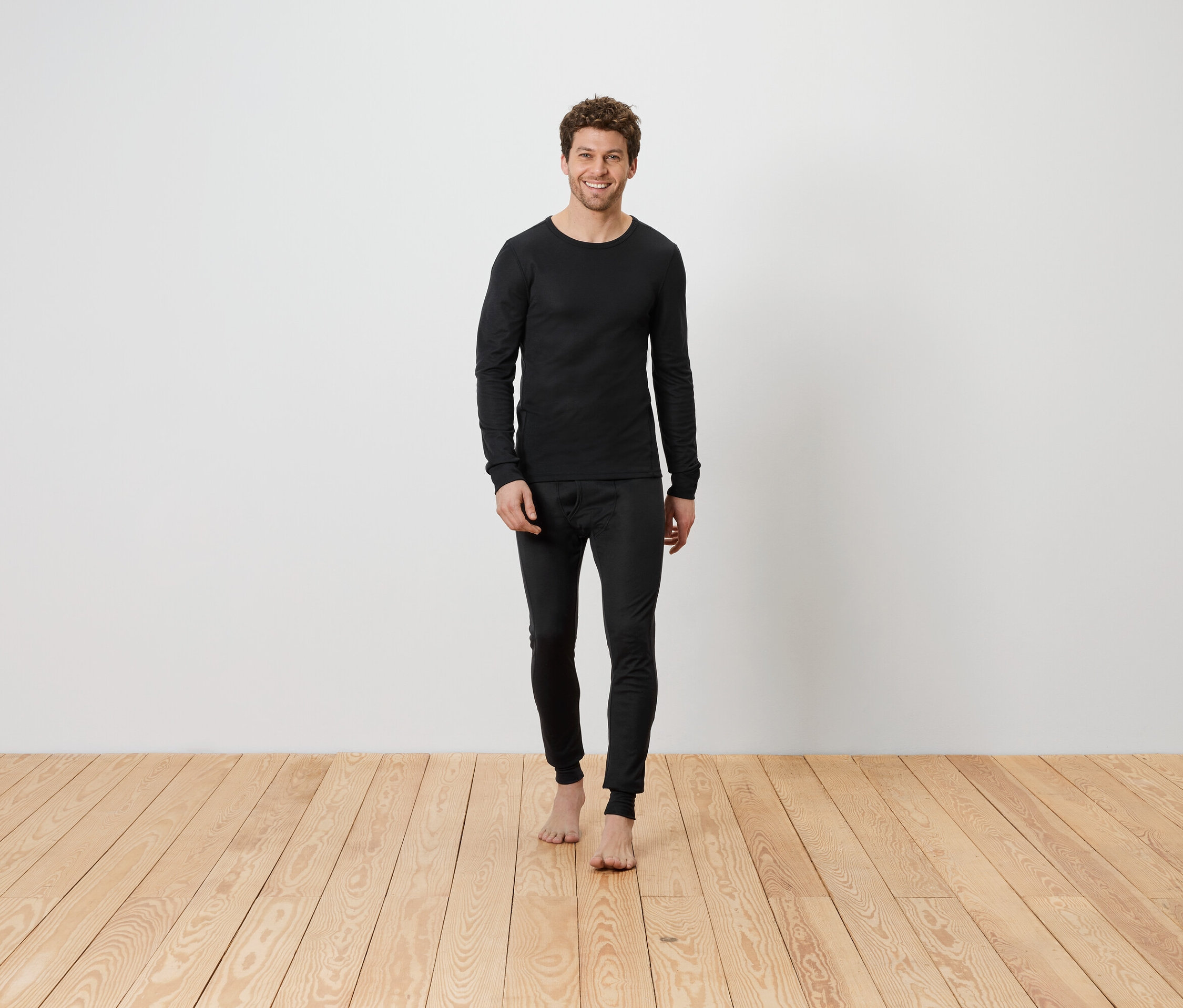 Ein Mann in schwarzer Thermounterwäsche steht barfuß auf einem Holzboden vor einer weißen Wand.