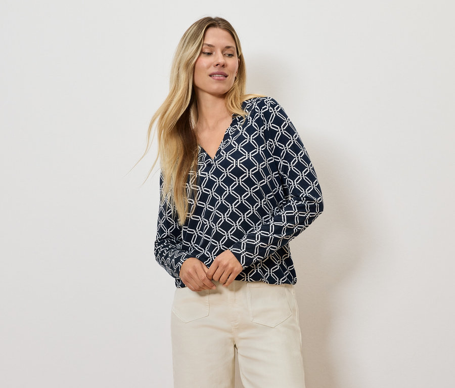 Blonde Frau posiert in einer geometrisch gemusterten Bluse und hellen Hosen.