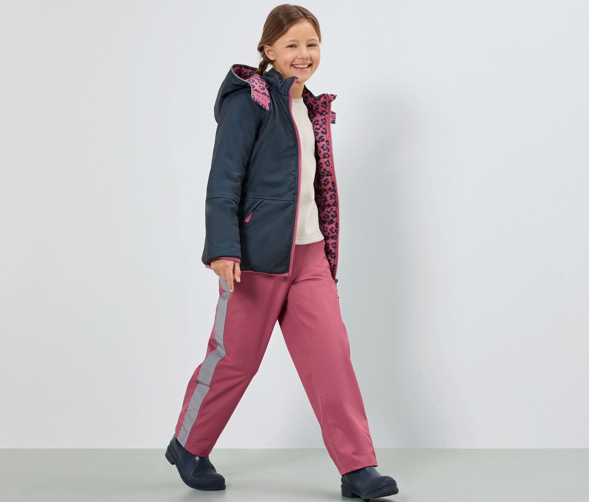 Lächelndes Mädchen geht und trägt eine dunkelblaue Jacke und eine rosa Kinder-Regenhose.