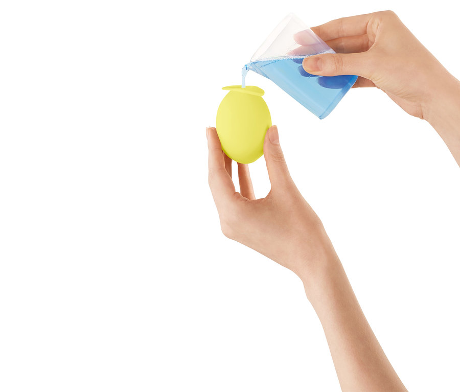 Hände gießen blaue Flüssigkeit aus einem Messbecher in ein gelbes, eiförmiges Gefäß.