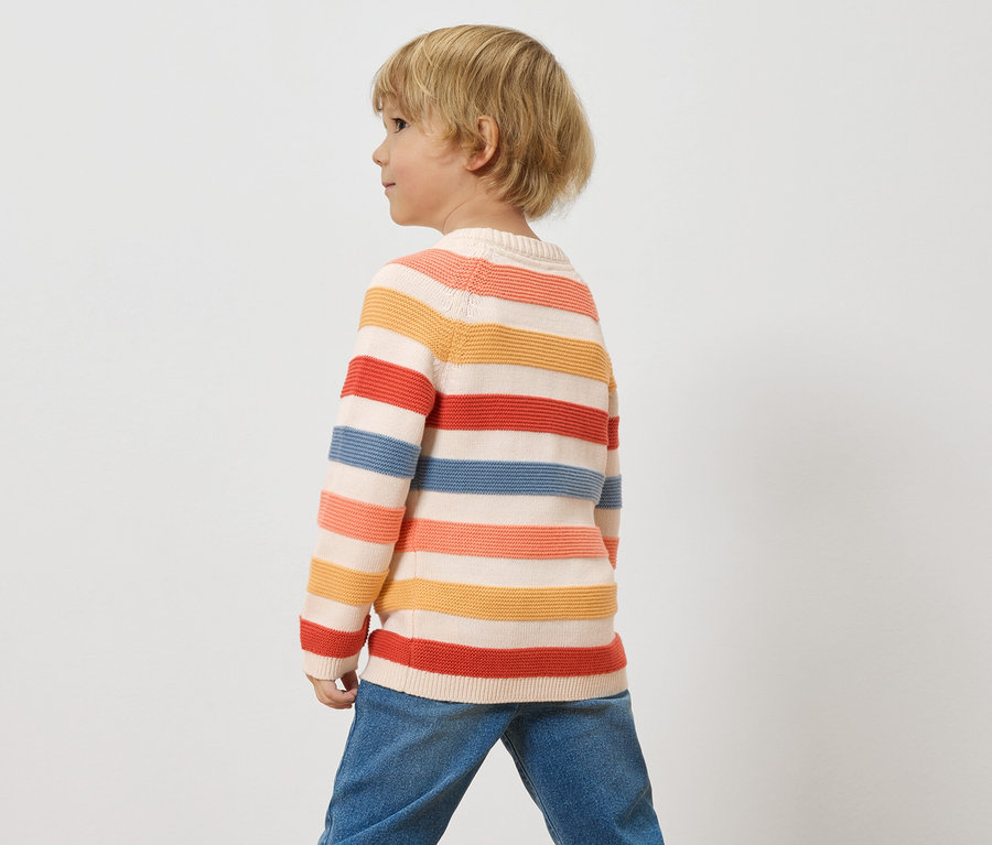 Ein Kind mit blonden Haaren trägt einen gestreiften Pullover und Jeans vor einem weißen Hintergrund.