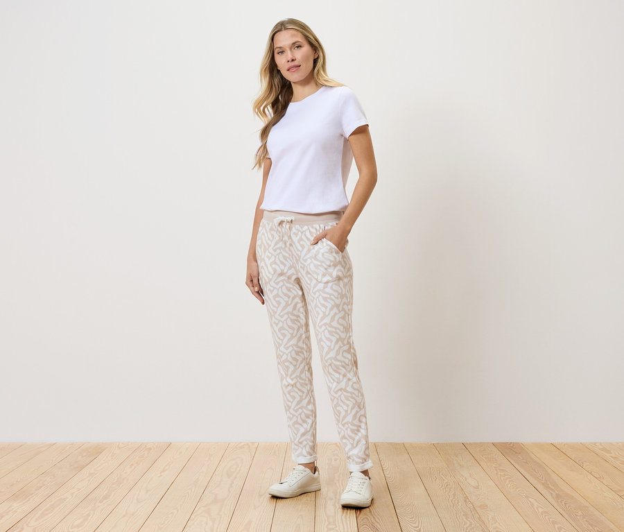 Frau steht vor einer Wand, trägt ein weißes T-Shirt, gemusterte Hose und weiße Turnschuhe.