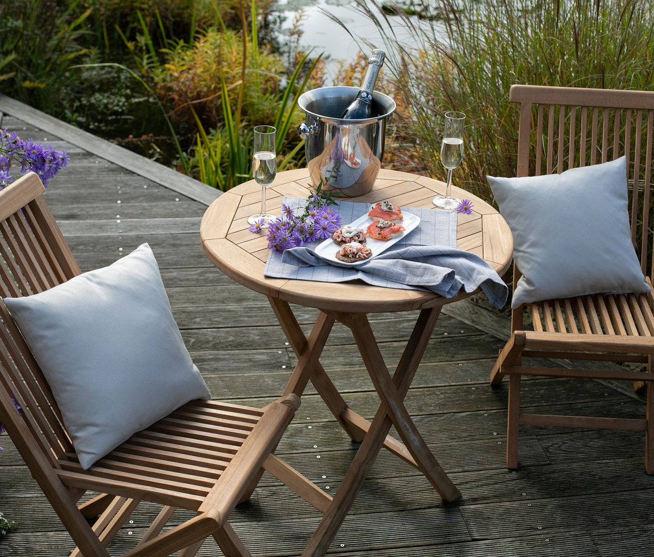 Ein runder Holztisch mit Sekt, Snacks und zwei Holzstühlen mit Kissen auf einer Holzterrasse.