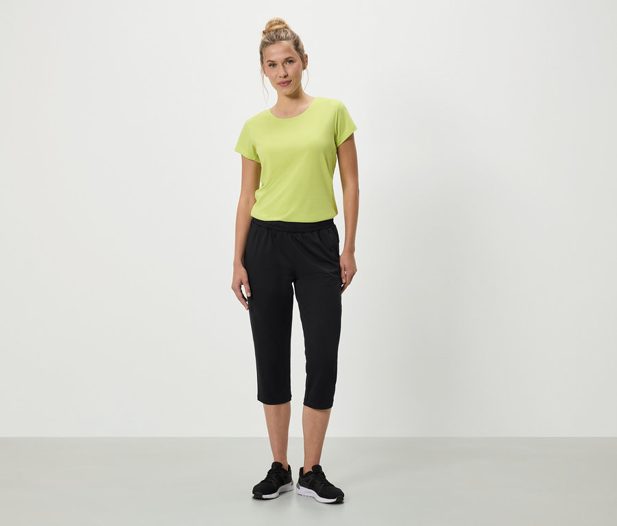 Frau steht mit grünem T-Shirt, schwarzen Dreiviertelhosen und schwarzen Turnschuhen vor einem weißen Hintergrund.