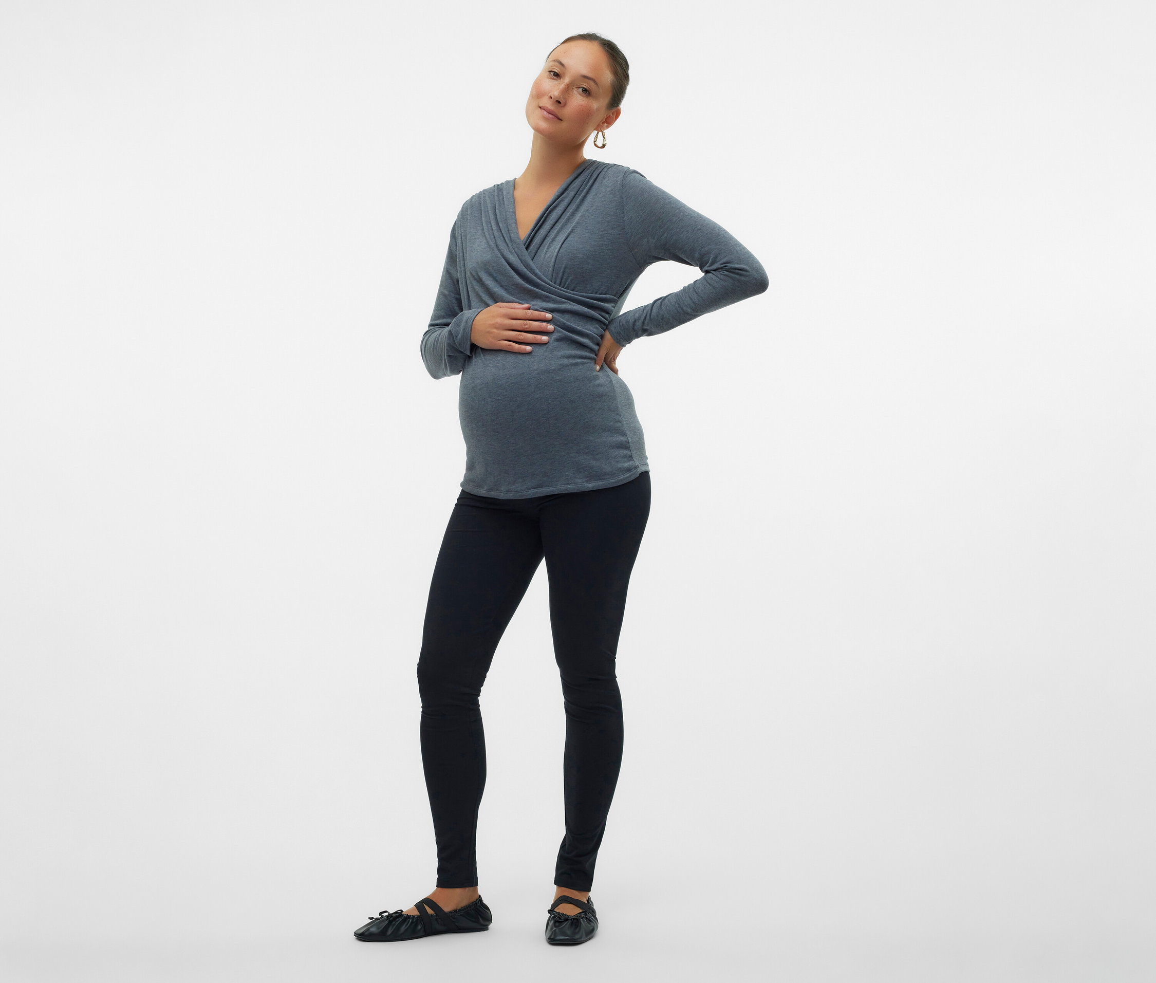 Schwangere Frau in grauem Wickeloberteil, schwarzen Leggings und Ballerinas posiert vor einem weißen Hintergrund.