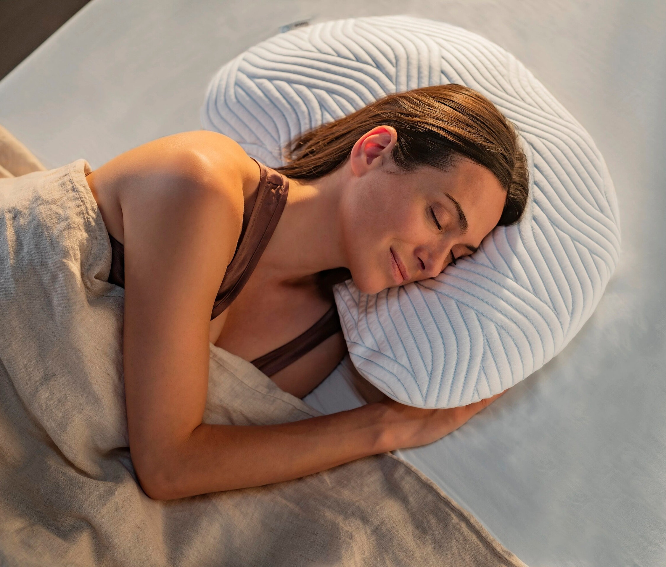 Frau schläft auf der Seite, auf einem weißen Bett unter einer beigen Decke und mit einem hellblauen, geformten Kissen.