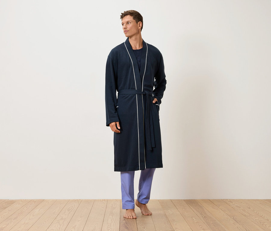 Ein Mann steht barfuß auf einem Holzboden, bekleidet mit einem marineblauen Morgenmantel mit weißer Paspelierung und blauer Hose.