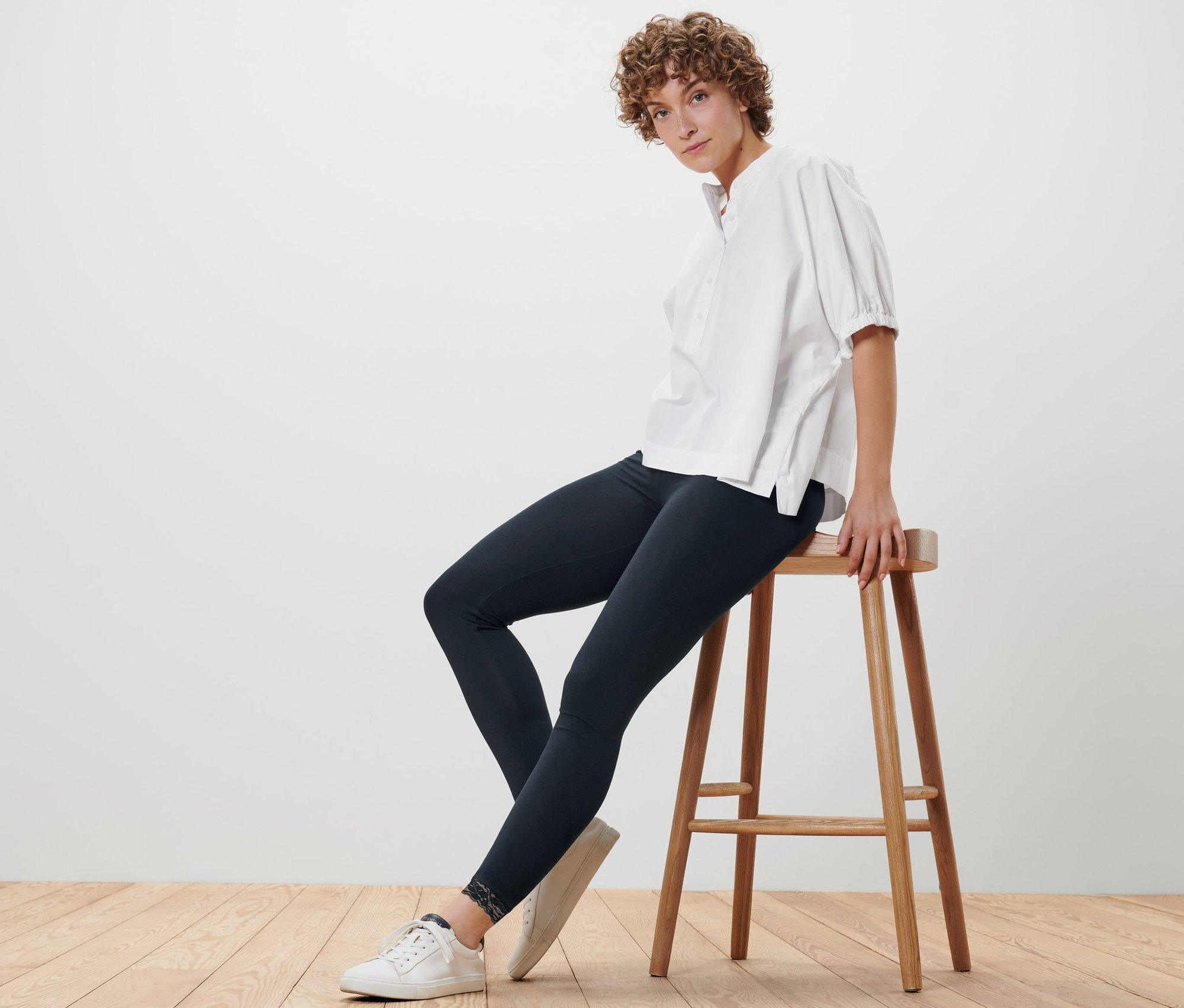 Frau sitzt auf einem Hocker und trägt schwarze Leggings mit Spitzenborte und eine weiße Bluse.