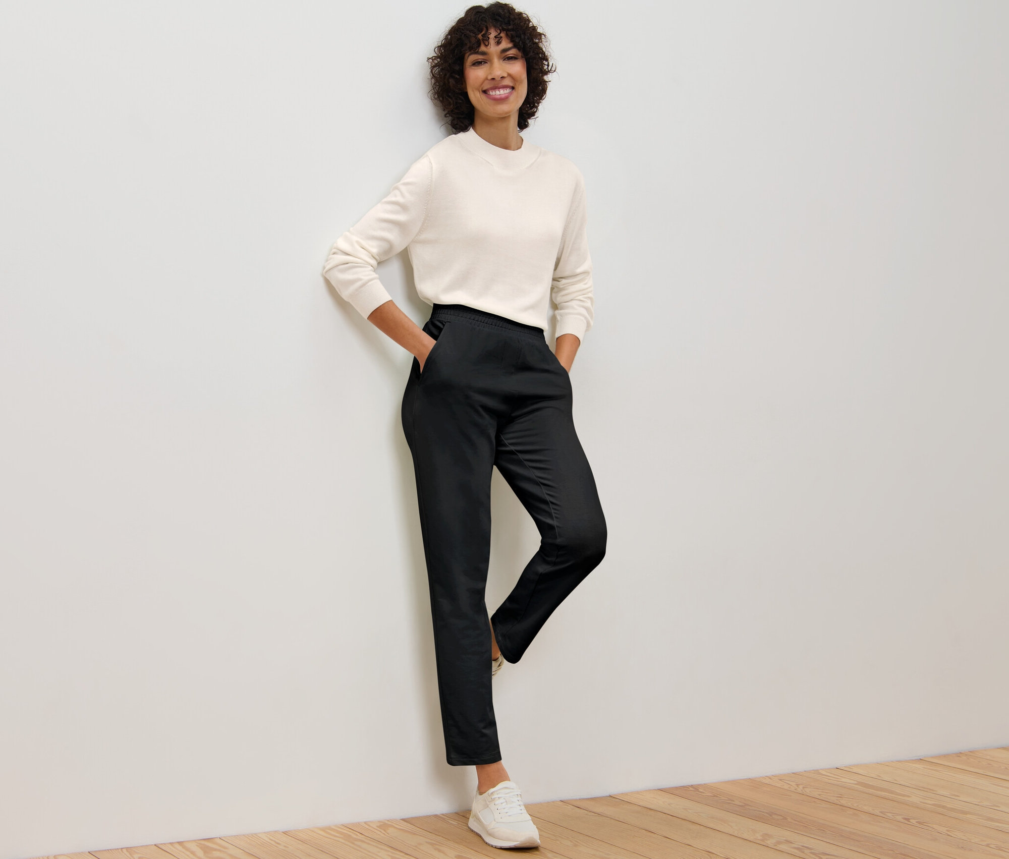 Eine Frau lehnt an einer Wand und trägt eine schwarze Joggpants und ein cremefarbenes Oberteil.