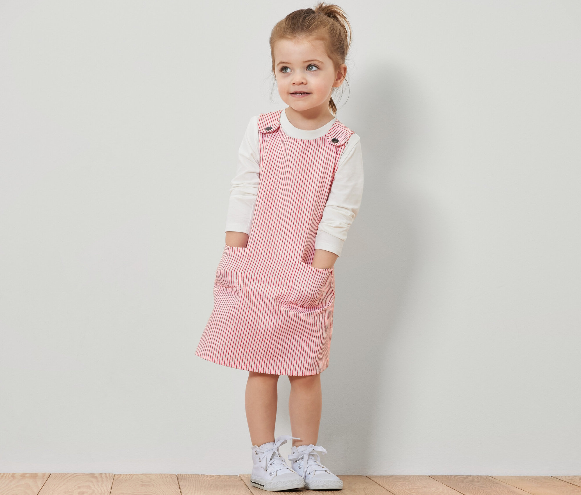 Mädchen trägt ein Kinder-Web-Latzkleid mit roten Streifen über einem weißen Langarmshirt.