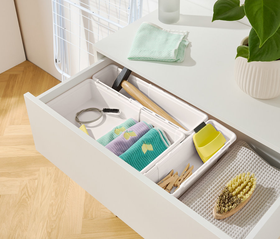Offene Schublade einer weißen Aufbewahrungseinheit mit verschiedenen Gegenständen, darunter Tücher, ein Hammer und eine Bürste.
