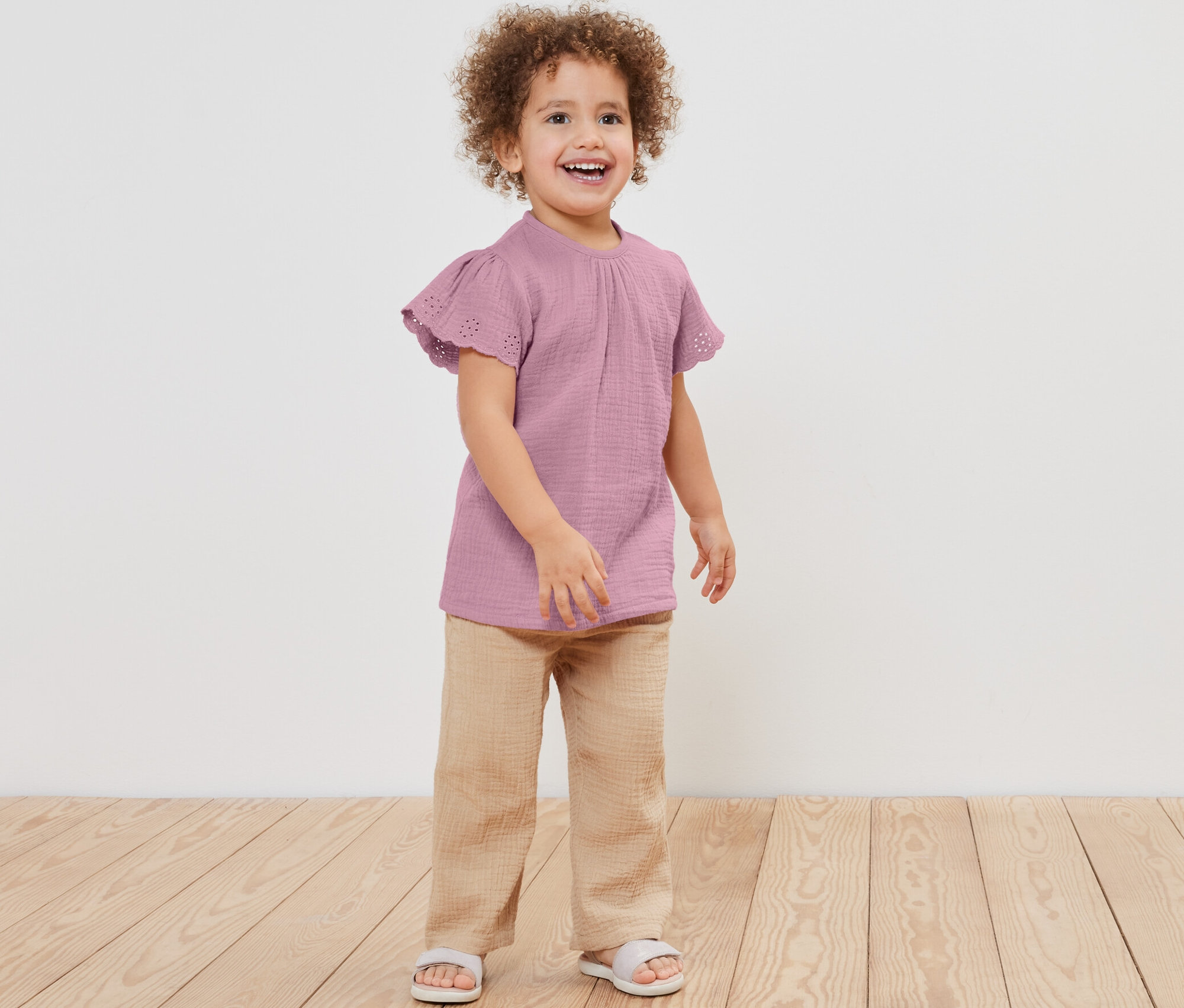 Lächelndes Mädchen posiert in Kinder-Musselin-Shirt, Hose und Sandalen.