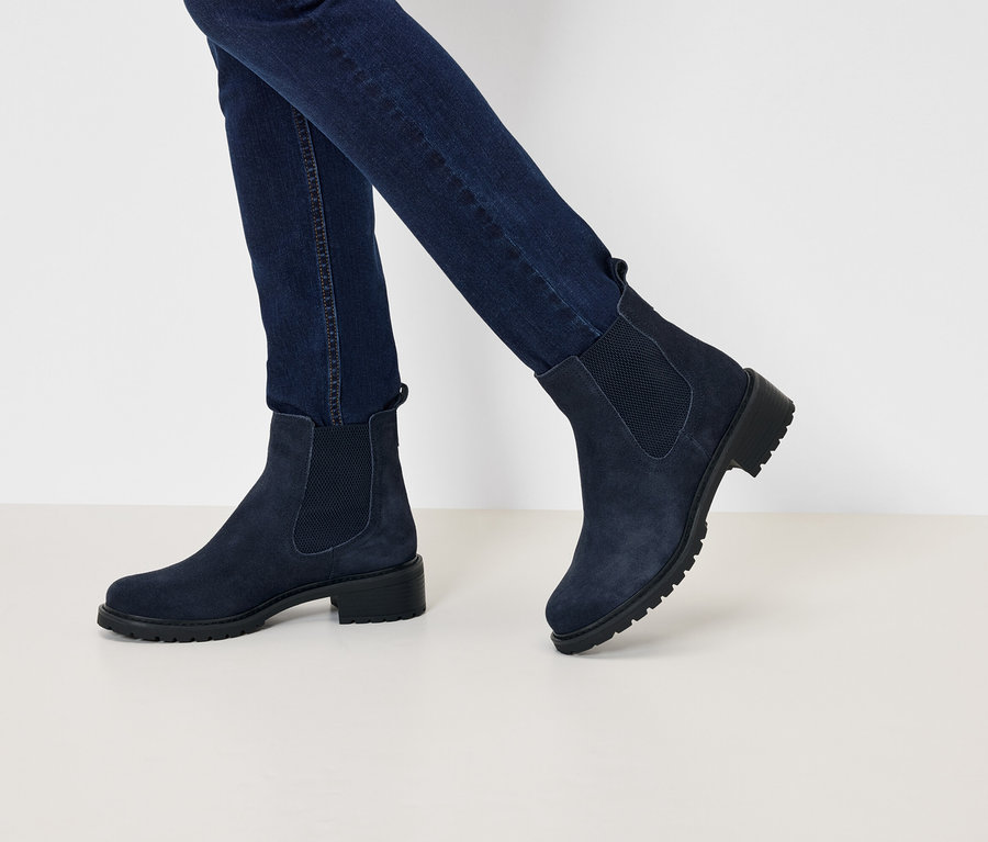 Beine in dunkelblauen Jeans und marineblauen Wildleder-Chelsea-Stiefeln mit niedrigem Absatz, die auf einer weißen Oberfläche laufen.