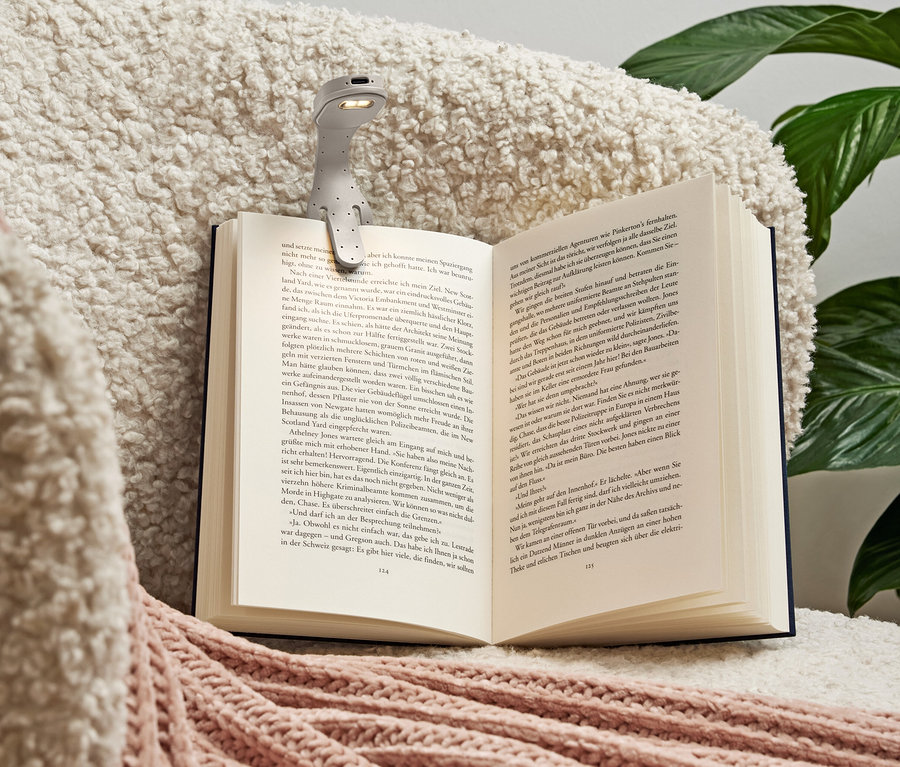 Eine graue Leselampe ist an ein aufgeschlagenes Buch geklemmt, das auf einer weichen, cremefarbenen Decke mit rosa Akzenten liegt.