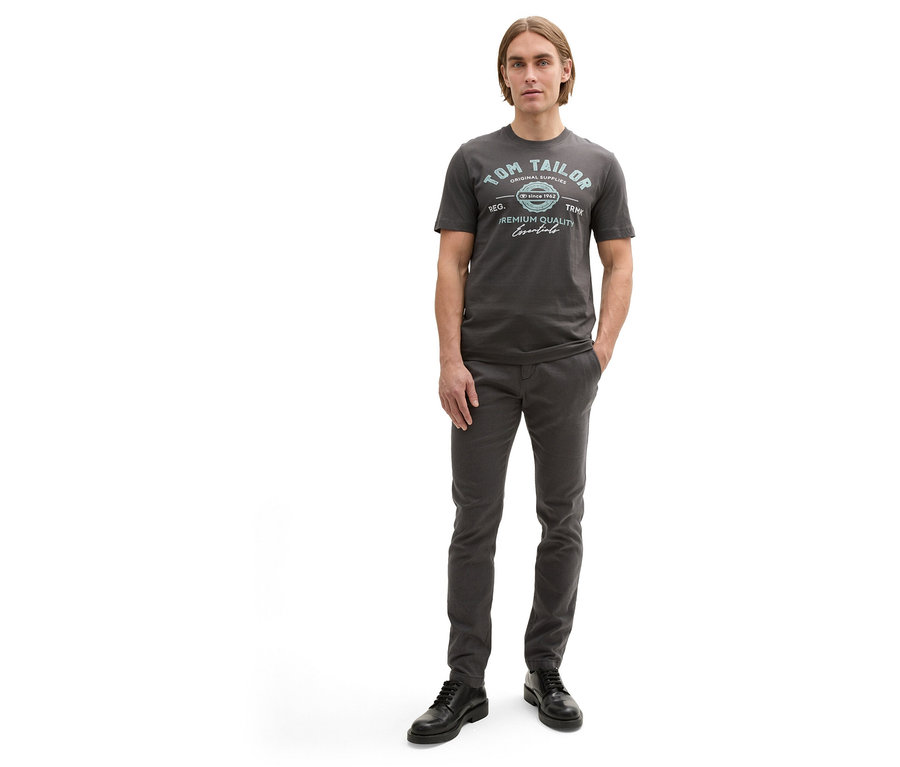 Mann in Hose und Tom Tailor T-Shirt mit Logo, Tarmac Grey, die Hände in den Hosentaschen.