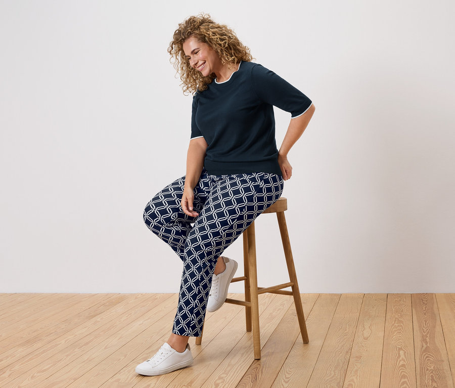 Frau mit lockigem Haar sitzt auf einem Hocker und trägt ein marineblaues Oberteil und gemusterte Hosen.