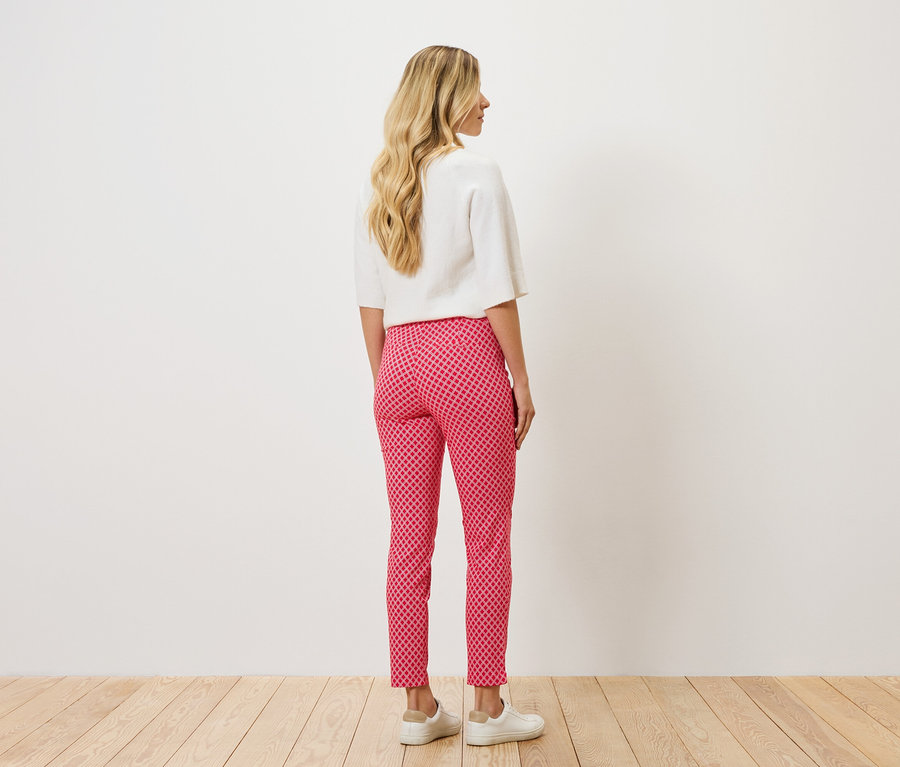 Rückansicht einer Frau mit blonden, gewellten Haaren, die eine weiße Bluse und eine rot-pink gemusterte Hose mit weißen Schuhen trägt.