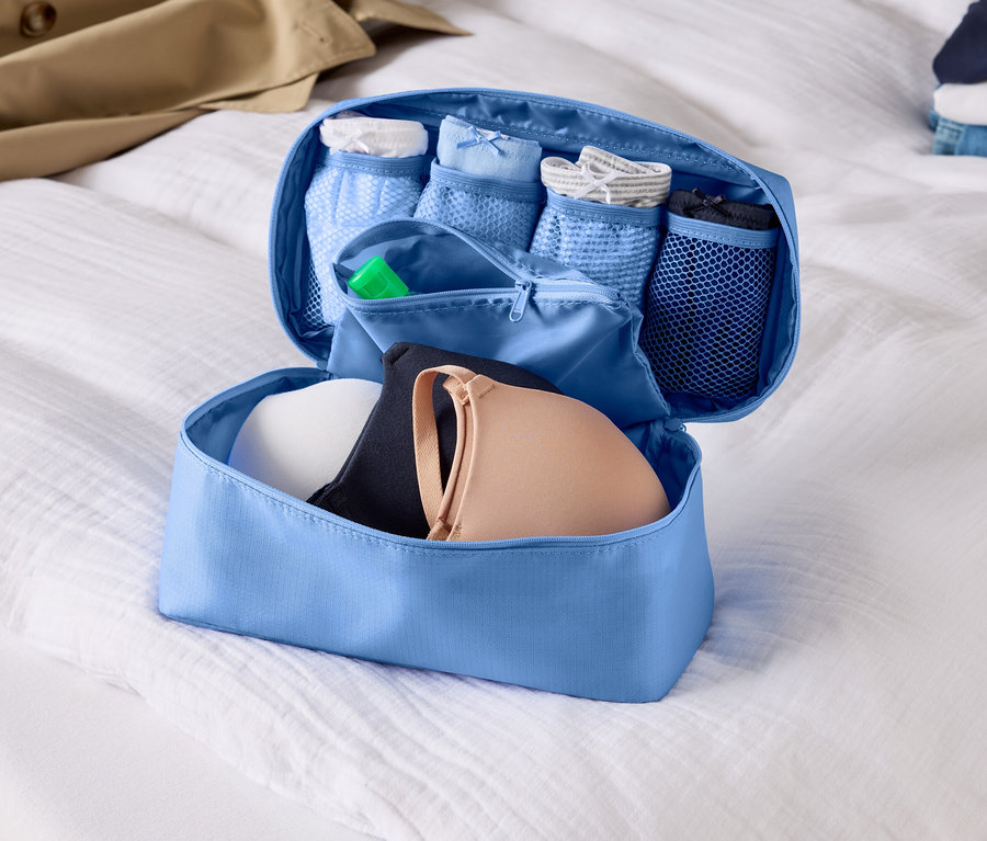 Offener, hellblauer Gepäckorganizer mit BHs und Unterwäsche auf einem weißen Bettlaken.