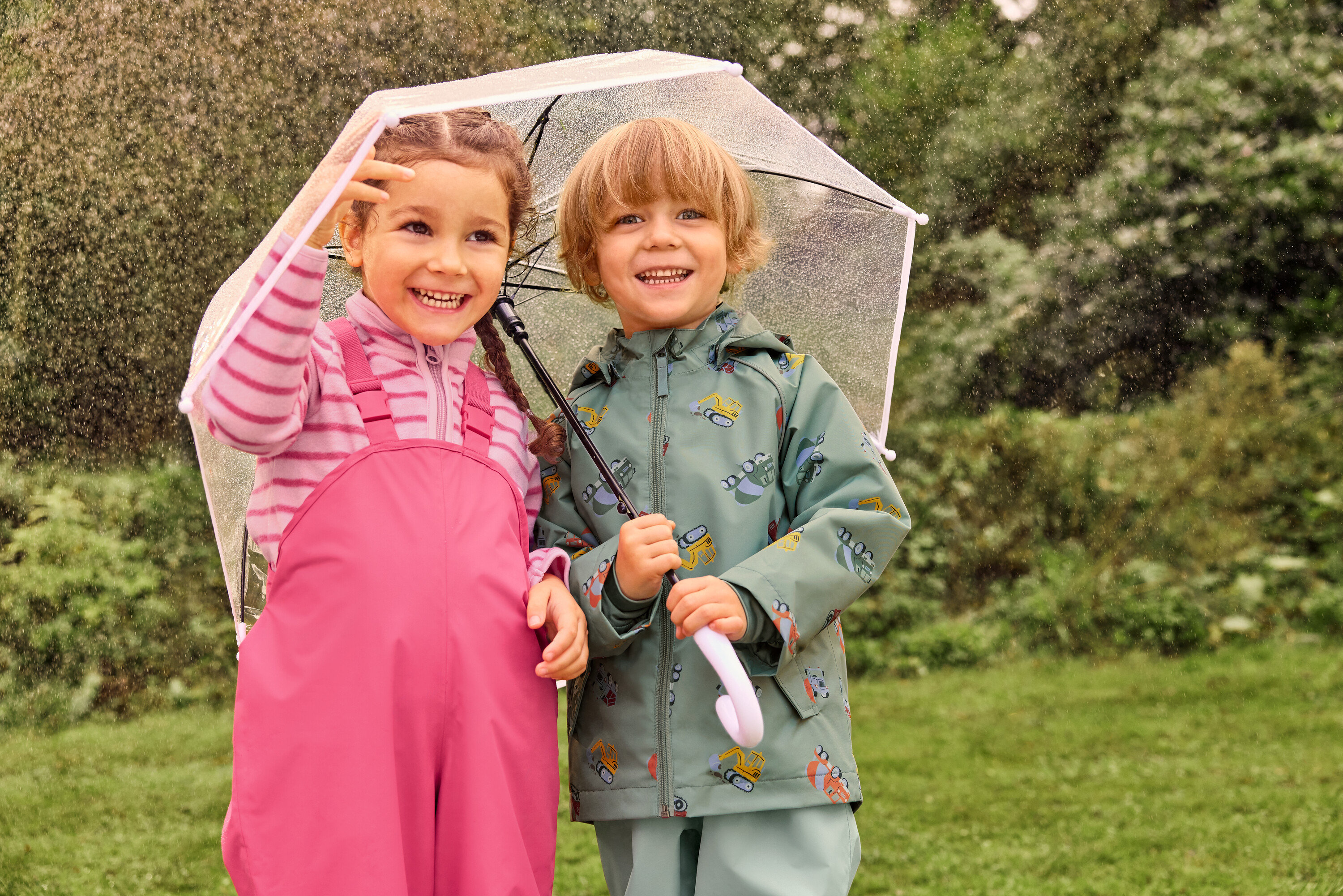 Zwei lachende Kinder, ein Mädchen in rosa Latzhose und ein Junge in einer grünen Jacke mit Bagger-Motiv, stehen unter einem durchsichtigen Regenschirm im Regen.