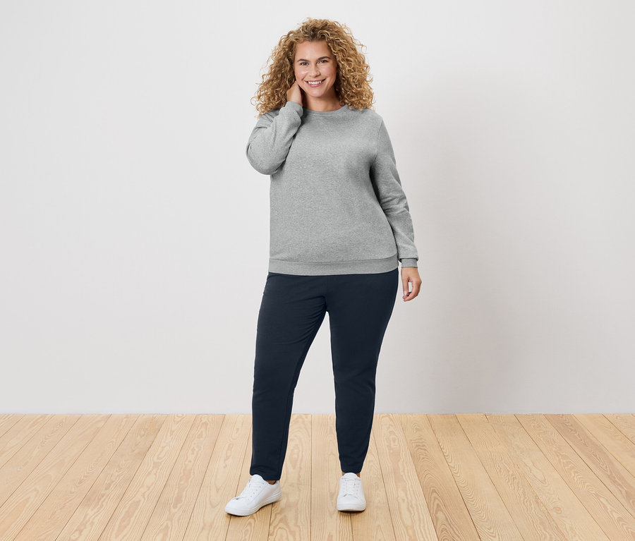 Frau mit lockigem Haar, die einen grauen Pullover und dunkelblaue Hosen trägt.