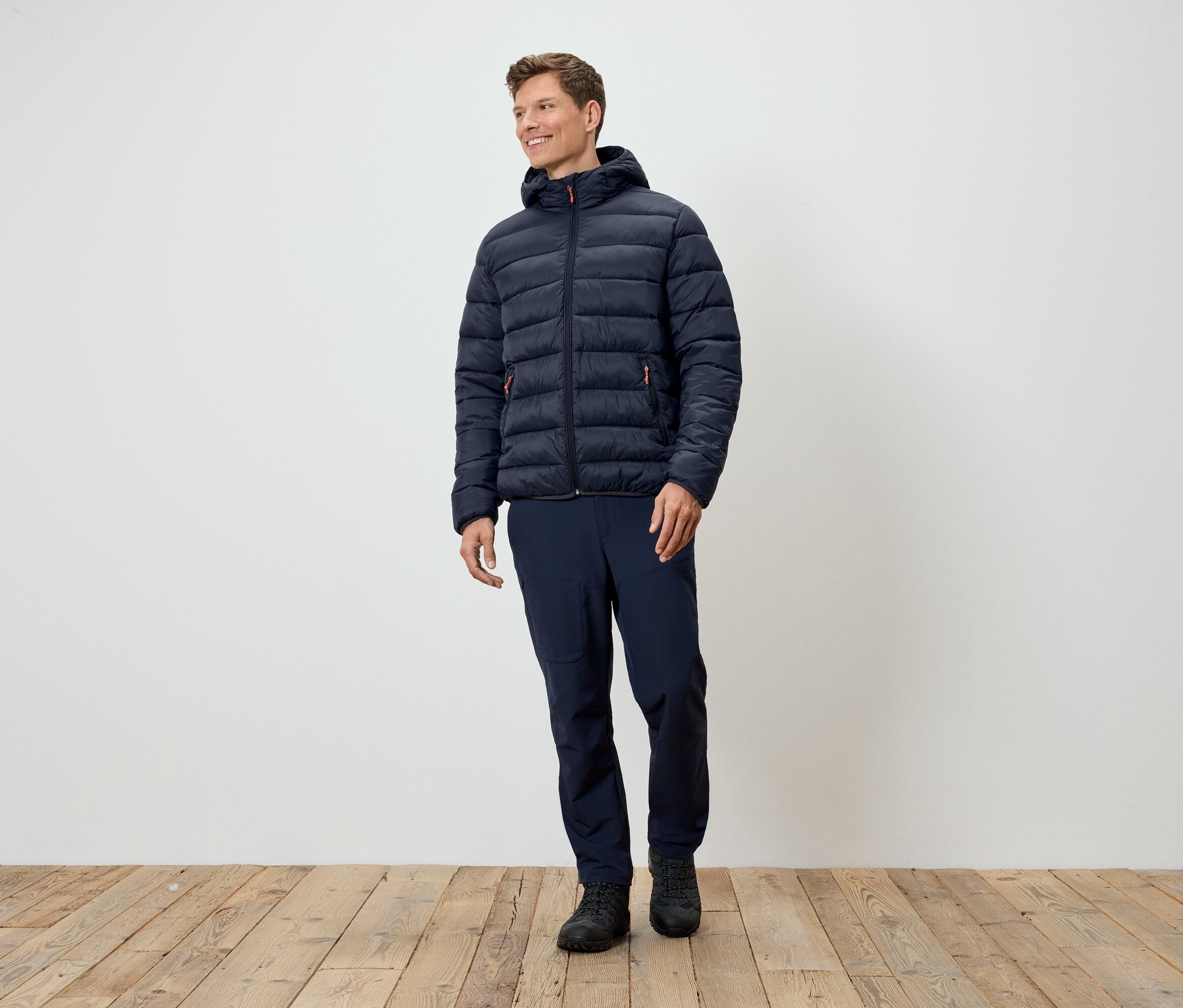 Ein Mann steht und trägt eine dunkelblaue Steppjacke, dunkelblaue Hosen und schwarze Schuhe.