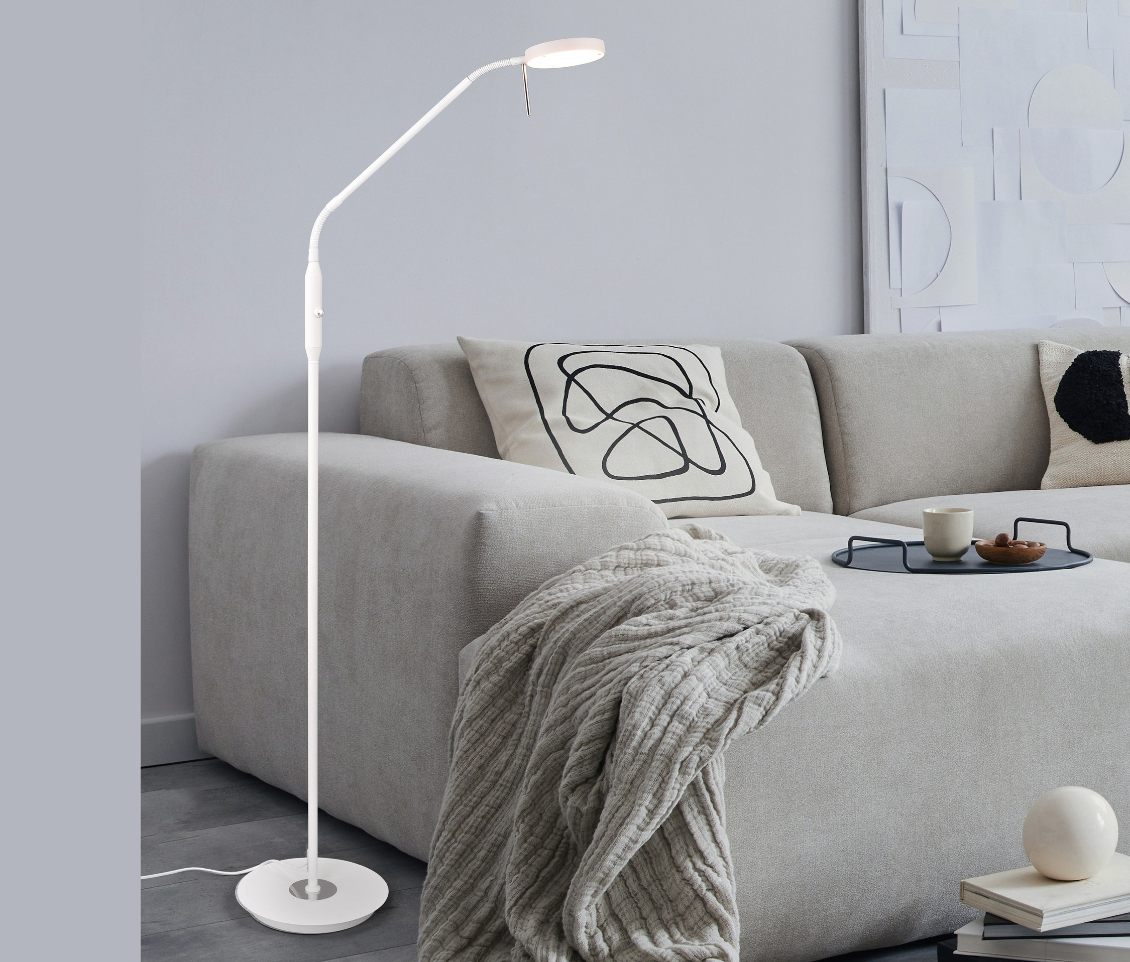 Weiße Trio LED-Stehleuchte »Monza« steht neben Sofa mit Decke und Kissen.