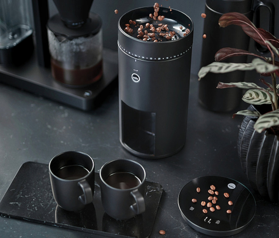 Eine Hand schüttet Kaffeebohnen in eine schwarze Wilfa Kaffeemühle »Svart Uniform+« neben zwei Tassen Kaffee.