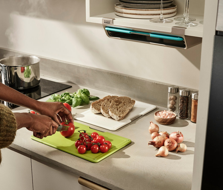 Eine Frau schneidet eine Tomate auf einem grünen Joseph Joseph Schneidebrett. Auf der Arbeitsfläche liegen Zwiebeln, Brot und Gewürze.