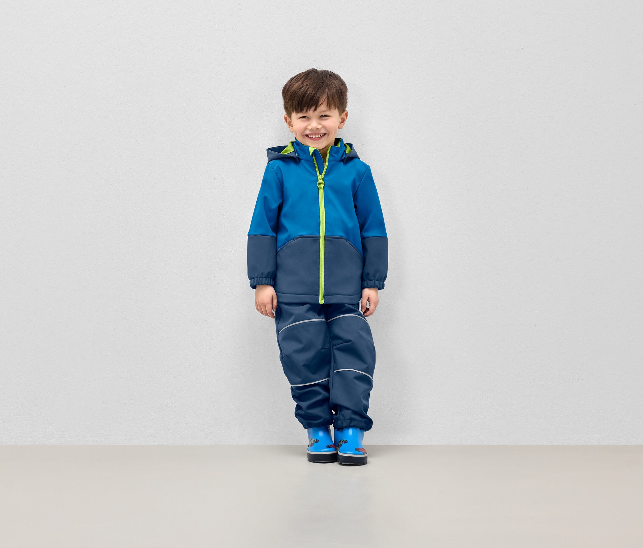 Lächelnder Junge posiert in blauer Jacke mit Kapuze und dunkelblauer Hose mit reflektierenden Streifen.