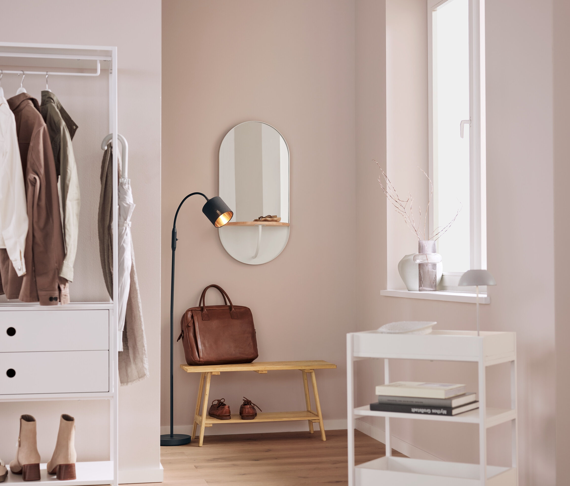 In einem Interieur befinden sich ein weißes Regal mit Kleidung, ein ovaler Spiegel, eine braune Ledertasche auf einer Bank, eine schwarze Lampe und ein weißer Rollwagen.