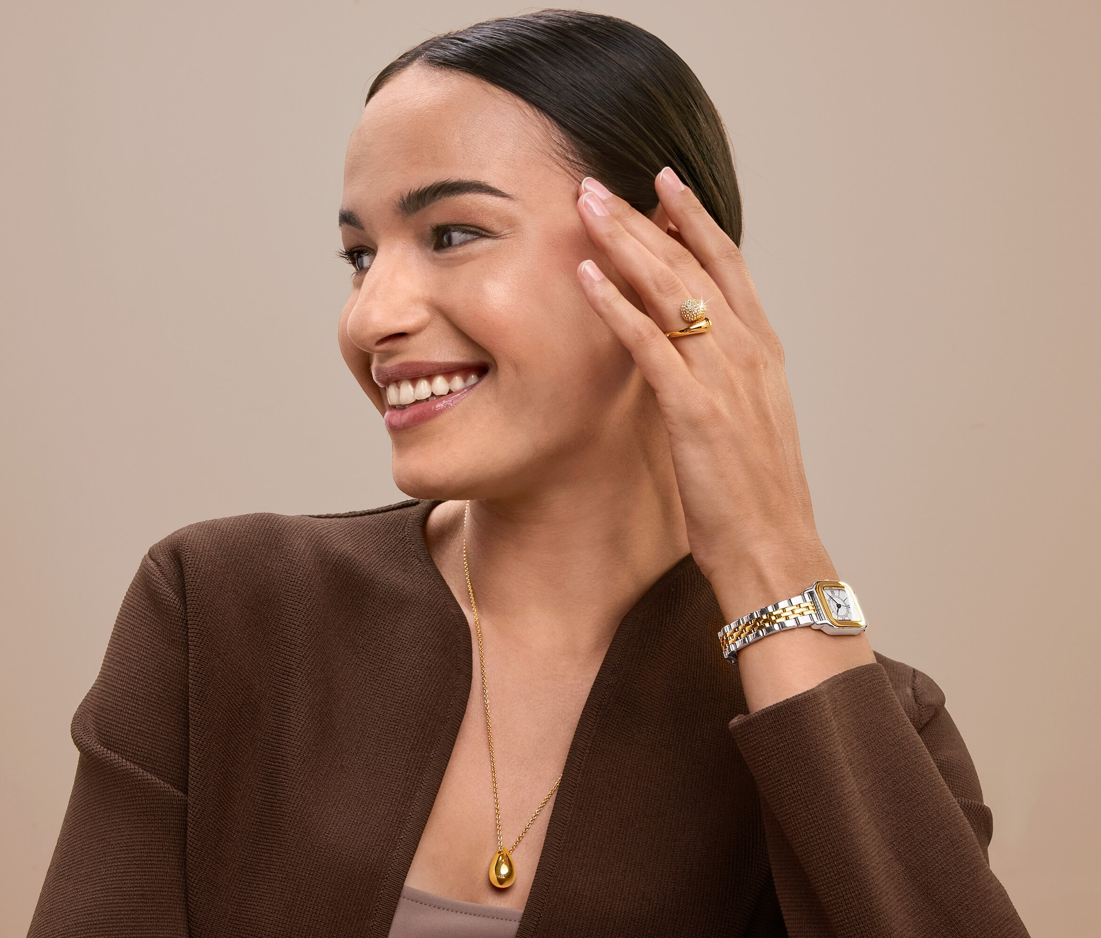 Lächelnde Frau berührt ihr Gesicht und trägt einen goldenen Ring, eine Uhr und eine Halskette.