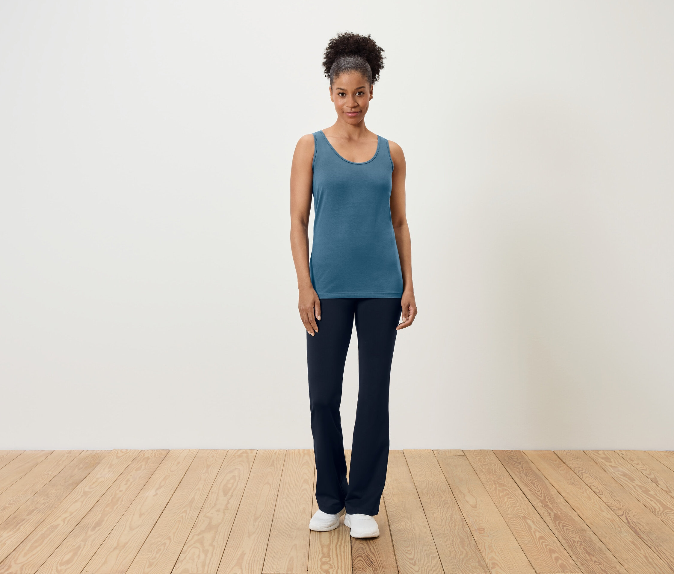 Eine Frau steht auf einem Holzboden und trägt ein blaues Tanktop und dunkelblaue Hosen.