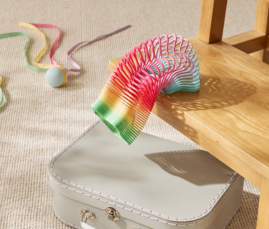 Eine regenbogenfarbene Spielzeugspirale liegt teilweise auf einem beigen Koffer unter einem Holzstuhl.