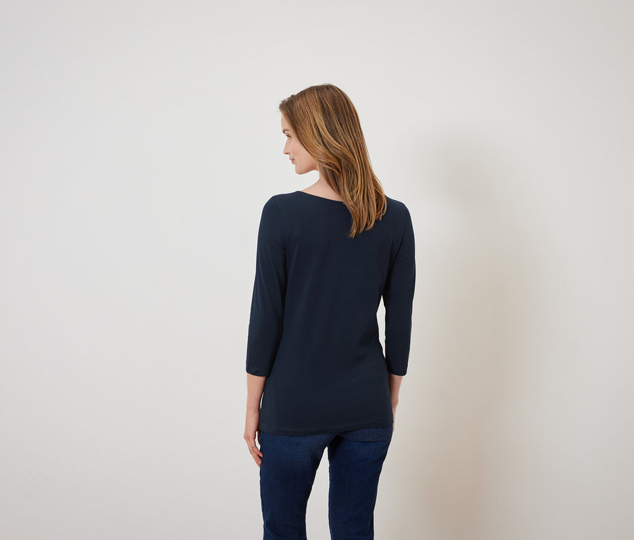 Eine Frau steht mit dem Rücken zur Kamera und trägt ein dunkelblaues Dreiviertelarmshirt und Jeans.