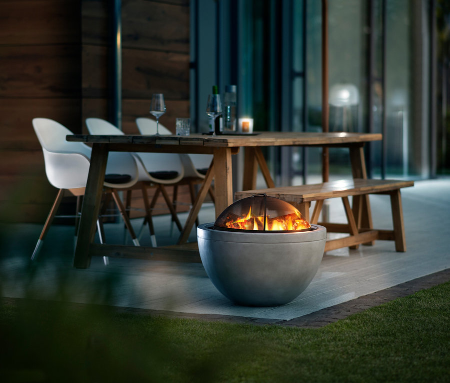 Feuer brennt in einer runden Feuerschale aus Beton vor einem Holztisch mit Gläsern und Stühlen im Freien.